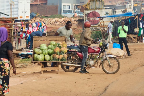 People on a motorcycle next to a wooden box full of watermelon and pineapple, Entebbe, Uganda