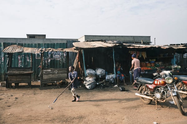 Child playing with a roller by a store front and fruit stand, Uganda